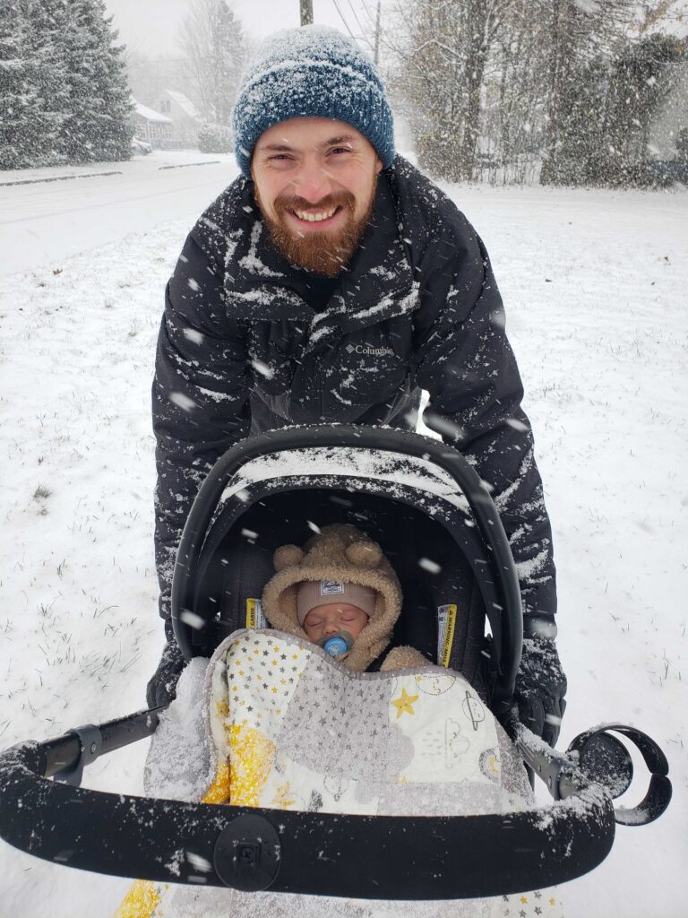 A father holding an infant in a carrier smiling during a snowfall 