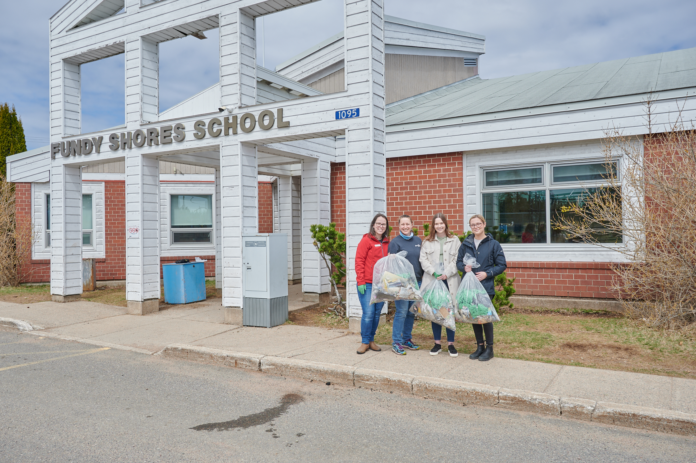The Gaia Project staff standing in front of Fundy Shores School, holding bags of trash collected from school grounds 