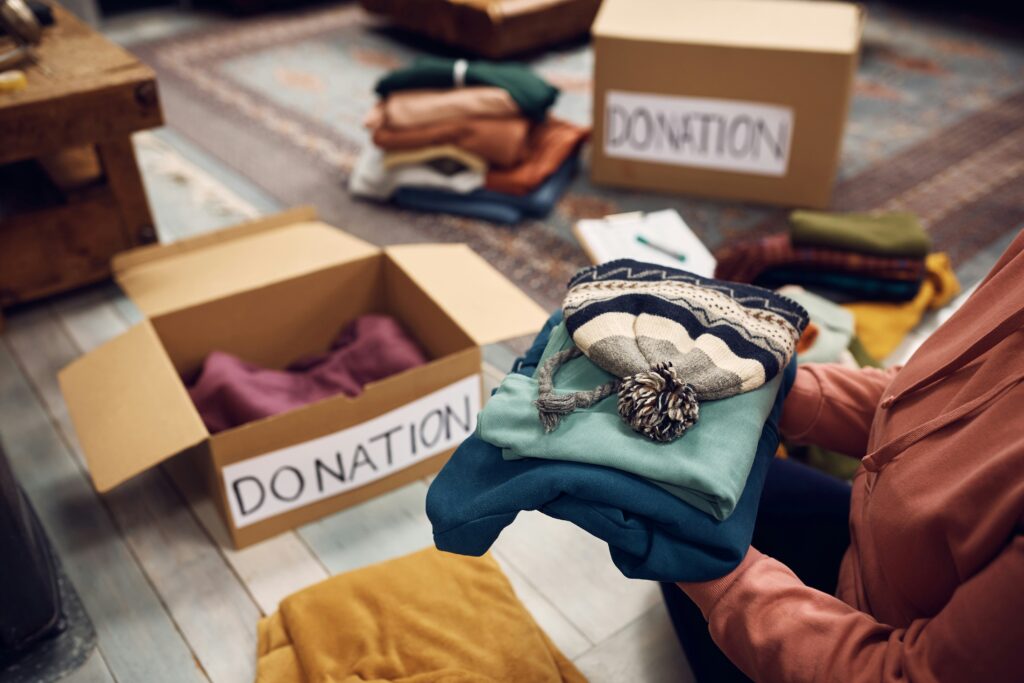A person holds a folder pile of winter clothes, a donation box sits on the floor 